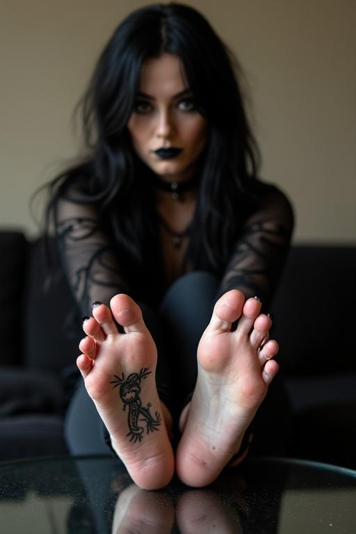 Mature goth woman with long black hair and black lips. Shows tattooed soles on bare feet. Sitting on couch with feet on glass table. Wearing goth clothing.