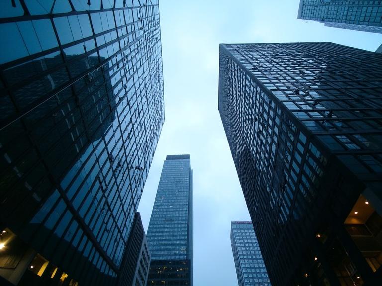 Photograph of a low angle cityscape. Looking up at tall modern skyscrapers. Reflective glass facades. Dark, sleek buildings against a blue sky. Some clouds in the background. Geometric shapes and sharp angles. Urban architecture with symmetry. Soft lighting and a cool color palette. Windows illuminated with lights on inside. Modern design in architectural photography. Perspective highlighted and vertical lines emphasized. Setting is metropolitan and urban. No people present. Focused on high-rise buildings and glass facades.