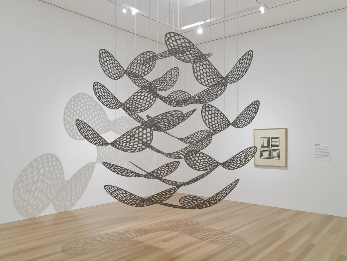 Large suspended mobile sculpture features multiple tiers of twisted wire forms resembling birds in flight. Each element is balanced for movement with air currents. Shadows shift on gallery walls. Style inspired by Alexander Calder and Naum Gabo. Kinetic wire sculpture demonstrates engineering poetry.