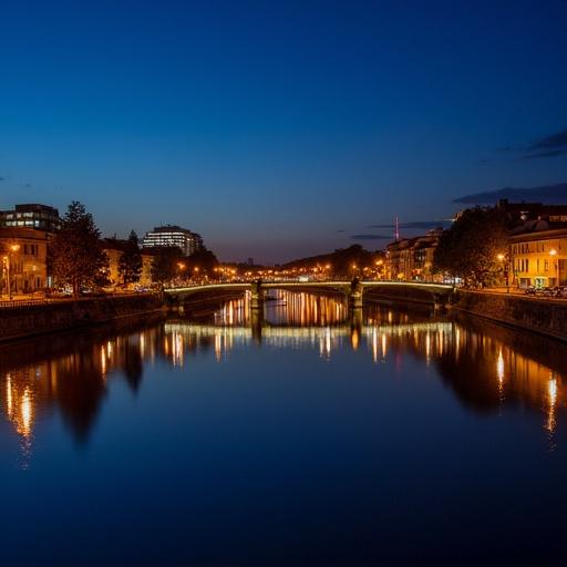 Mainz waterfront at night showing a calm river reflecting city lights. The illuminated bridge is prominent. The sky is deep blue, creating a tranquil atmosphere. Buildings line the riverbanks.