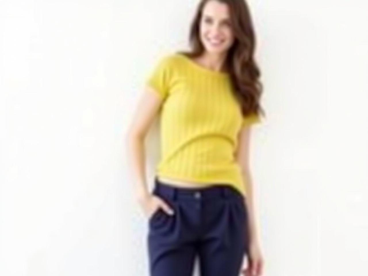 The image features a person standing against a plain white background. They are wearing a fitted yellow ribbed top that has short sleeves. The individual also has on navy blue high-waisted pants, which are pleated. Their hair is long and styled, cascading down their back. The person is posing with one hand in their pocket and looking slightly off to the side. The overall vibe is professional and casual.