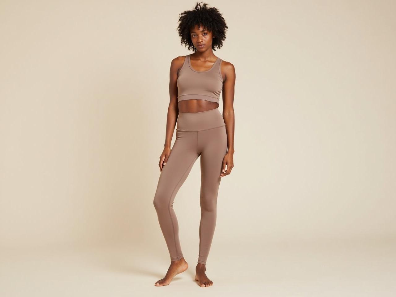 The image depicts a person standing in a neutral-toned background. They are wearing a matching set consisting of a fitted crop top and high-waisted leggings. The outfit is in a light brown or taupe color. The person has curly hair and is barefoot. The overall look is sporty and stylish, showcasing a modern athleisure outfit.