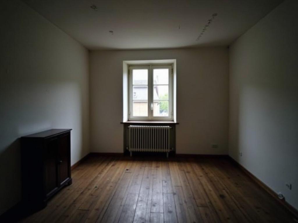 The image shows a dimly lit room with white walls and a wooden floor. There is a window with shutters on the left side, and a radiator is located beneath it. A small dark cabinet can be seen on the left wall. The atmosphere feels quiet and empty, suggesting an unused or unfurnished space. The light appears soft, creating shadows that add to the sense of emptiness in the room.