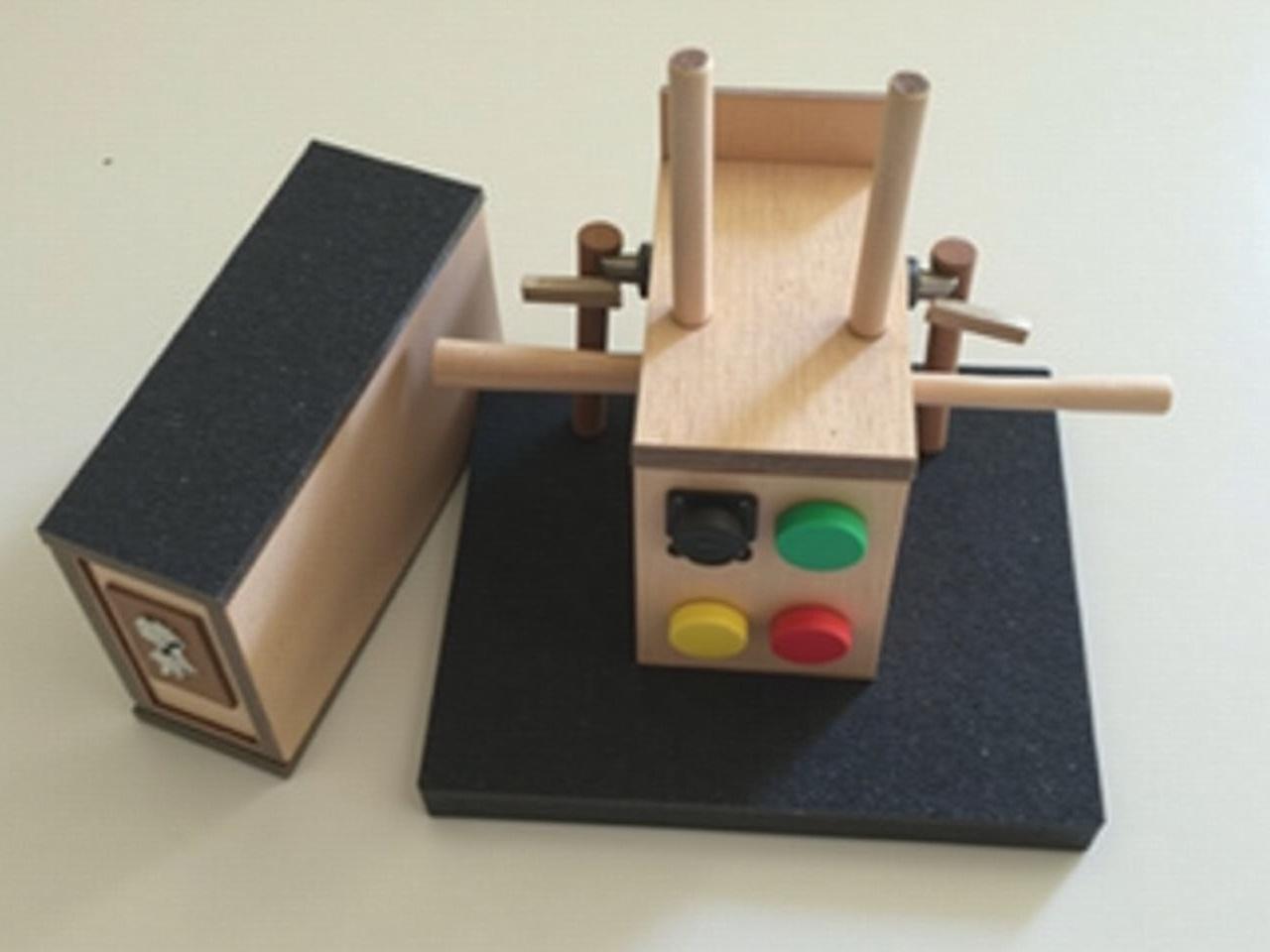 This image shows a small, handcrafted model, likely made from various materials including wood and black foam. It features a rectangular base with a flat top, possibly representing a piece of machinery or equipment. There are two cylindrical sticks placed horizontally on top, which could symbolize handles or levers. On the front, there are two colored buttons, one red and one green, suggesting a function or control mechanism. The overall design appears to be simplistic, aimed at demonstrating a concept rather than being a fully functional device.