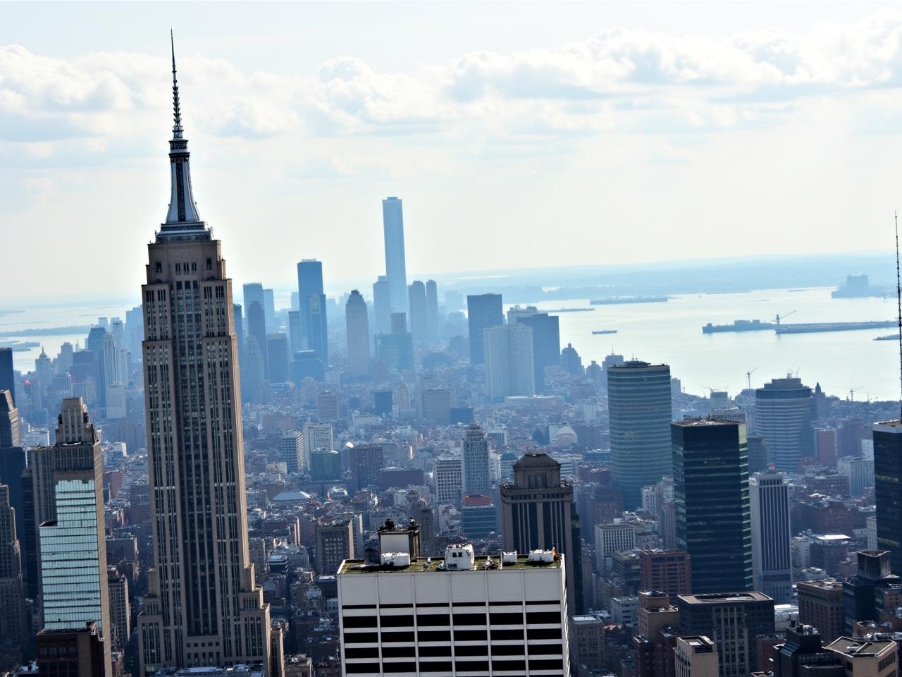 The image features a skyline view of a bustling city. In the foreground, a tall, iconic building with a distinctive spire stands out. Surrounding it are various other skyscrapers, creating a dense cityscape. There is a hazy atmosphere in the background, lending a soft focus to the distant buildings. The scene suggests a blend of historical and modern architecture, with clouds partially covering the sky. The water can be seen in the background, adding a serene element to the otherwise vibrant city setting.