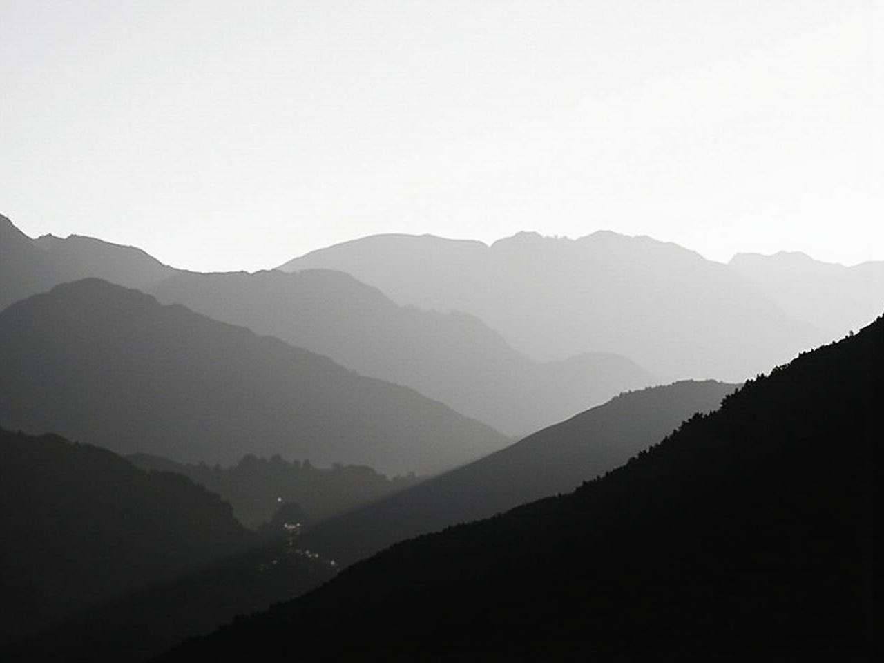 This image features a black and white silhouette of mountains. The foreground showcases layered mountain shapes with varying heights and contours. Soft light creates a serene atmosphere, gradually fading from the foreground to the background. The use of stark contrasts highlights the shapes while eliminating color distractions. This artwork evokes a sense of tranquility and the beauty of nature's simplicity.