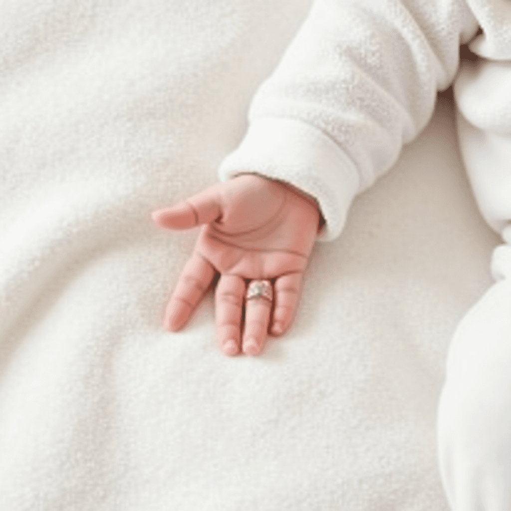 A baby's hand adorned with a delicate ring rests on a white blanket.