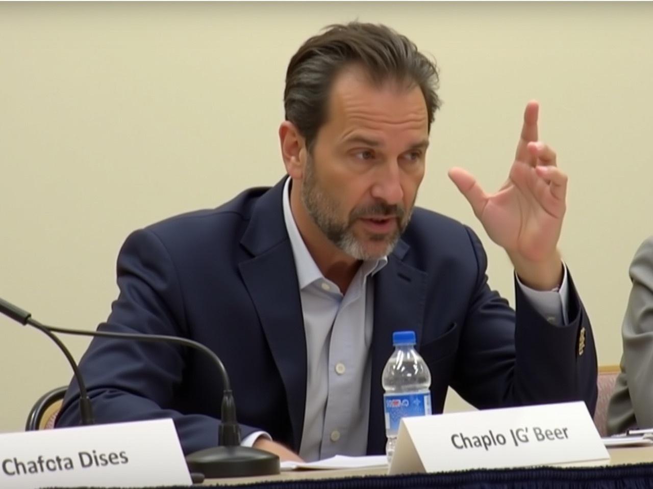 The image shows a man sitting at a table, partially obscured by a blurred face. He is wearing a navy blue blazer over a light-colored shirt. On the table, there is a microphone and a bottle of water. The setting appears to be a formal discussion or panel event. The man is gesturing with his hand, possibly emphasizing a point during his speech. The background shows a plain wall, suggesting a conference room or similar venue.