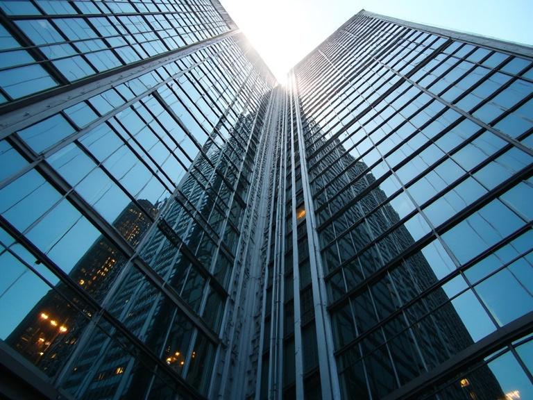 Angled view upwards of a glass skyscraper. Skyscraper reflects nearby buildings. Harsh midday sun creates reflections and shadows. Features repeating window patterns and metal beams.