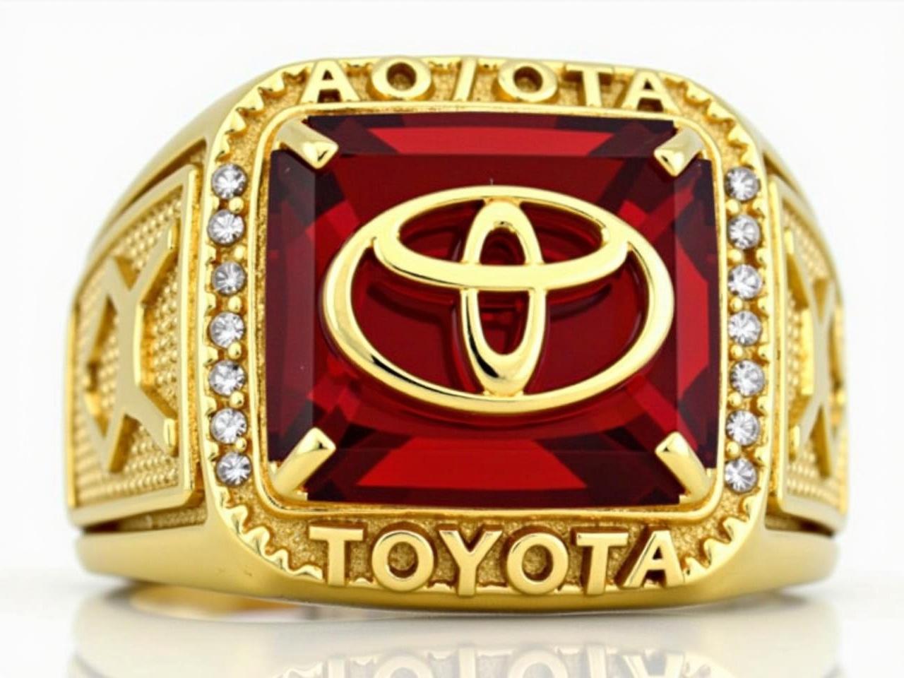 The image features a large, golden ring designed prominently with the Toyota logo. The surface of the ring has a red stone at its center, which showcases the logo in gold. Around the red stone, there are small diamond-like embellishments that add to the ring's design. The band of the ring bears the name 'TOYOTA' engraved in bold letters, emphasizing the brand. The overall design appears to be luxurious and striking, fitting for a championship or special commemorative item.