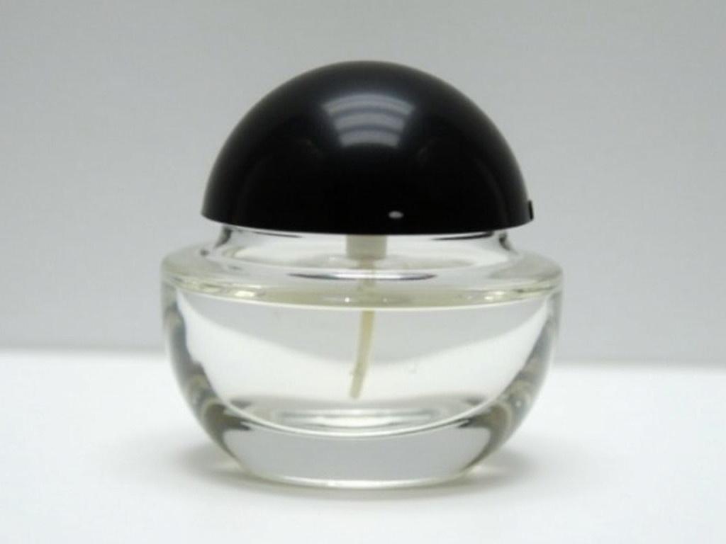 The image shows a small, clear glass container with a black dome-shaped lid. The container appears to be designed for holding a liquid, possibly a perfume or a similar substance. The glass is transparent, allowing visibility of the contents inside, which seem minimal. A label is visible on the side, although it is partially obscured. The focus is primarily on the sleek design of the container. The background is neutral, keeping the attention on the object.