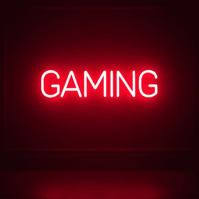 The image shows the word "GAMING" in neon lights. The letters are glowing in a bright light blue color. The background is dark, making the neon letters stand out. There is a slight red glow around the letters.