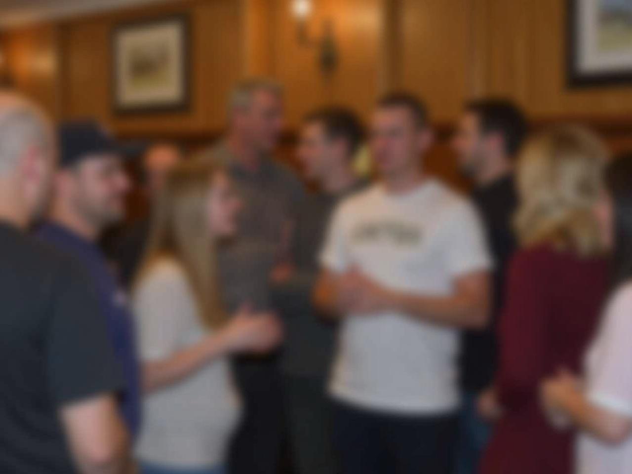 The image shows multiple people gathered indoors. They appear to be in a social setting, possibly a restaurant or bar. The background features wooden panels and a framed picture on the wall, indicating a cozy or traditional atmosphere. The individuals are engaged with each other, suggesting conversation or social interaction. Some faces are blurred to ensure privacy.