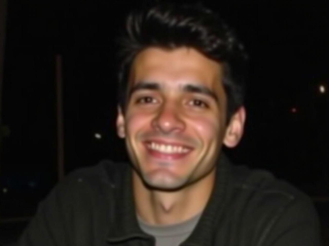 The image is a close-up portrait of a young man. He has dark hair styled casually and is wearing a dark jacket over what appears to be a light sweater. His expression is relaxed and thoughtful, with a slight smile. The background is blurred and dark, focusing the attention on his face. The lighting highlights his features, creating a soft contrast. He looks directly at the camera, inviting connection.