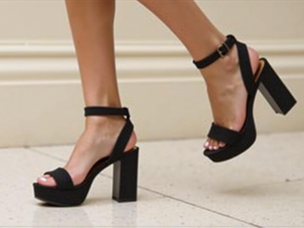 The image shows a close-up of a person's legs wearing stylish black heeled sandals. The sandals have a simple yet elegant design, featuring two straps across the foot. The legs are positioned as if the person is in motion, with the left foot slightly lifted, showcasing the heel of the sandal. The background is slightly blurred, which helps to focus on the feet and sandals. There is a neutral-colored floor beneath the feet, providing contrast to the black sandals.