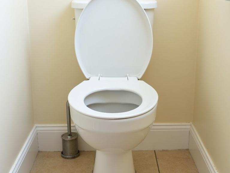 Photo shows a clean white toilet in a small bathroom. Lid and seat are up. Beige tiled floor, white baseboards, and cream-colored walls. Low angle view highlights the toilet bowl. Natural light provides bright even lighting. Simple and minimalistic setting with no water in the bowl. Metal flush pipe is visible on the left side.