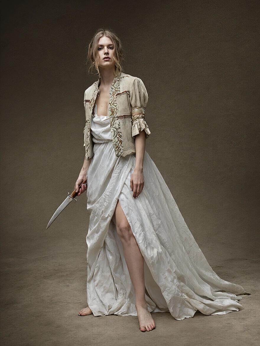 Woman standing barefoot holding a knife. She wears flowing white dress and a fitted jacket. Expression suggests strength and mystery. The background is neutral and textured.
